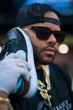 Tulpi wearing a black cap and sunglasses, holding a sneaker beside his face.