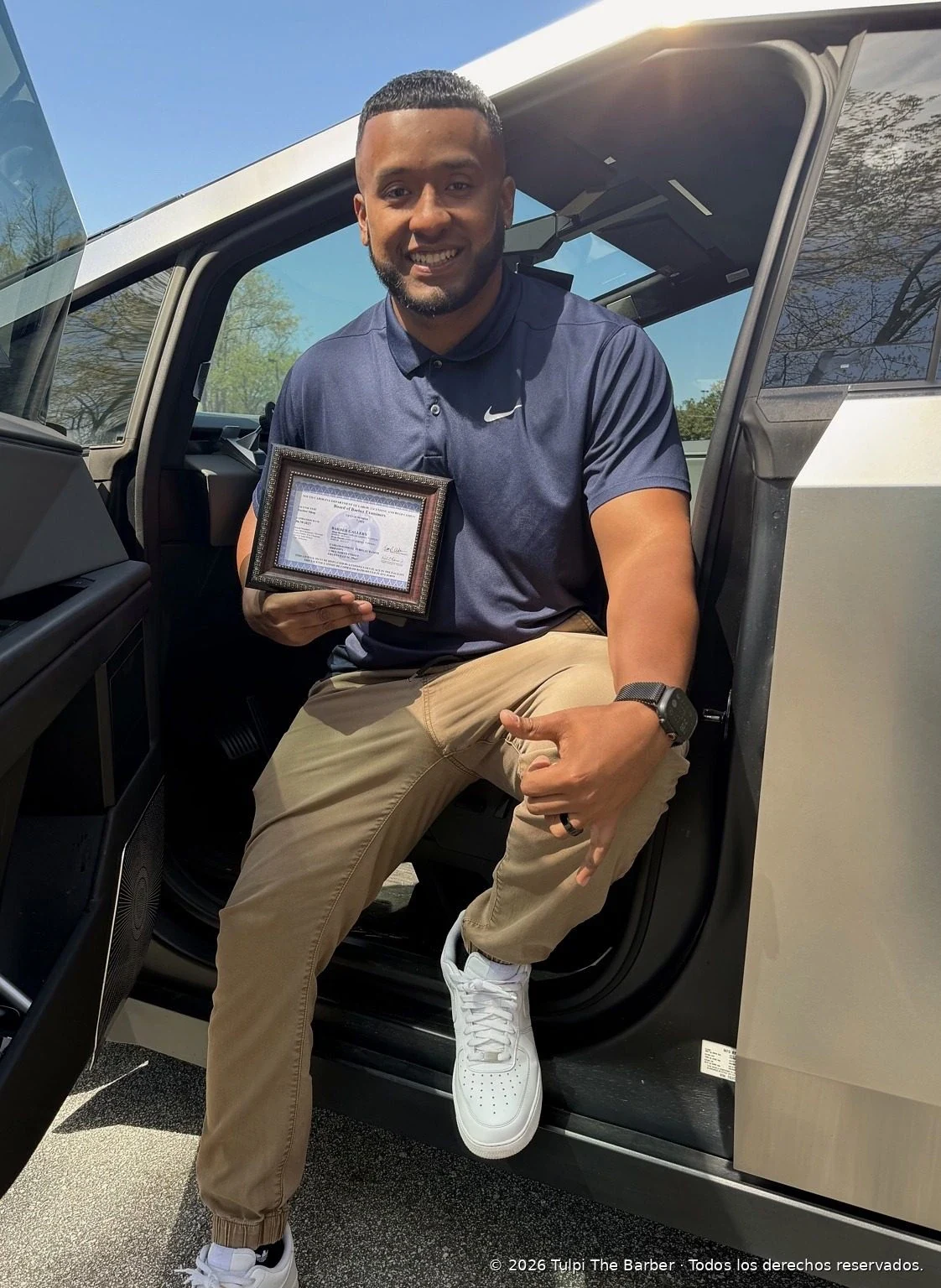 A Tulpi The Barber student smiling beside a vehicle while holding his framed barber license.