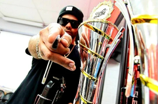 Tulpi wearing a black cap and sunglasses, pointing toward the camera beside his trophies.