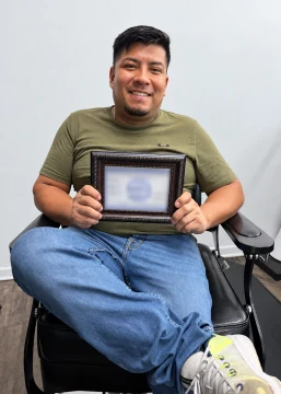 Mateo, wearing an olive-brown shirt, smiles while holding a framed document with its information blurred for privacy.