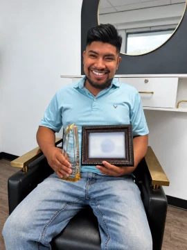 Luis, wearing a blue shirt, smiles while holding a trophy and a framed document with its information blurred for privacy.